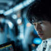 A close-up of a Formula 1 driver in a white racing suit, looking focused and deep in thought inside the team garage. The background is slightly blurred with other team members and technical equipment visible, creating a tense and concentrated atmosphere.