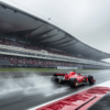 A red Ferrari Formula 1 car racing on a wet track under rainy conditions, leaving a trail of water spray behind. The race takes place at a large modern circuit with a grandstand full of spectators. In the background, the Shanghai skyline and the Oriental Pearl Tower are visible, indicating the race is held in China.