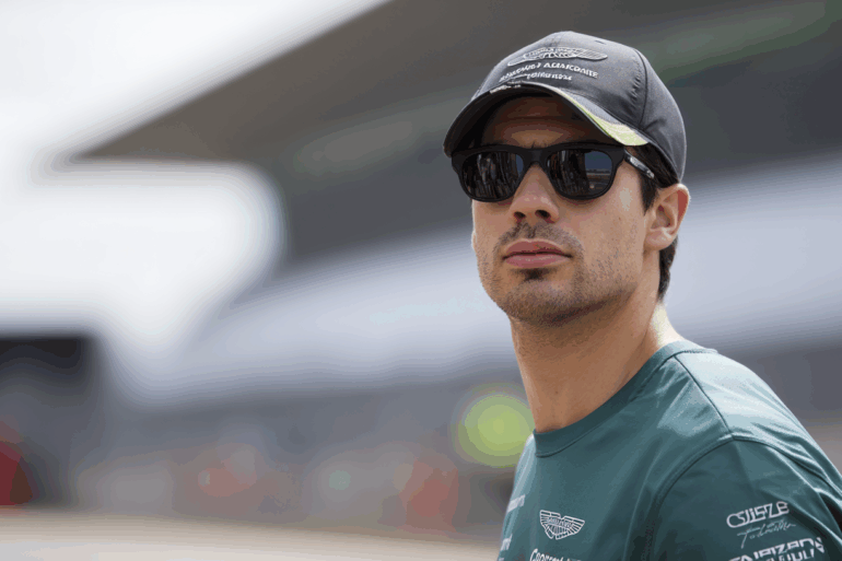 A Formula 1 driver in Aston Martin team gear wearing a branded cap and dark sunglasses, looking off to the side with a blurred race track structure in the background.