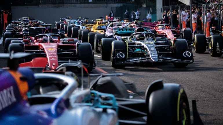 Multiple Formula 1 cars lined up on the starting grid at the Imola circuit, with a variety of team liveries visible, including Ferrari, Mercedes, and McLaren, under bright daylight with team members and spectators in the background.