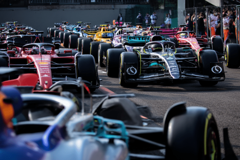 Multiple Formula 1 cars lined up on the starting grid at the Imola circuit, with a variety of team liveries visible, including Ferrari, Mercedes, and McLaren, under bright daylight with team members and spectators in the background.