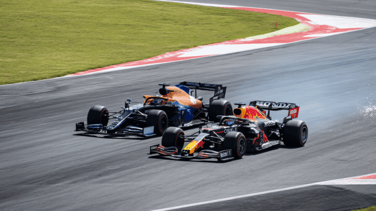 A Red Bull Formula 1 car races past a McLaren on a high-speed track corner, with visible sparks trailing behind the Red Bull car. The intense moment captures the action of a competitive overtake during a Formula 1 Grand Prix. ​