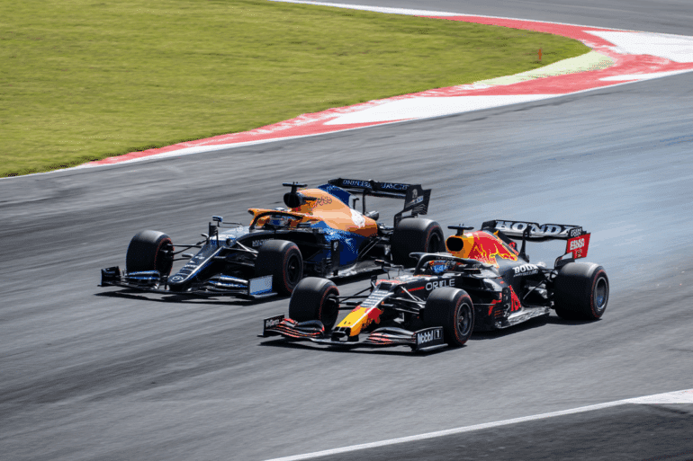 A Red Bull Formula 1 car races past a McLaren on a high-speed track corner, with visible sparks trailing behind the Red Bull car. The intense moment captures the action of a competitive overtake during a Formula 1 Grand Prix. ​