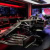 Ferrari Formula 1’s revolutionary new floor finally revealed, displayed on a technical stand inside the team’s garage. The matte black carbon fiber surface features intricate aerodynamic channels, sculpted fins, and hidden airflow innovations. Focused lighting highlights the engineering complexity, while digital screens in the background show schematics and airflow simulations. The scene feels secretive and cutting-edge, capturing the moment a closely guarded performance breakthrough is exposed.