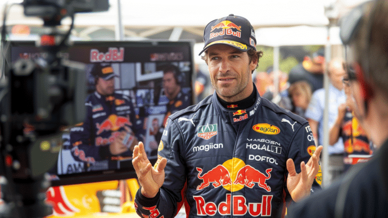 A Red Bull Formula 1 driver giving an interview in front of a camera crew. He is wearing a full Red Bull racing suit and cap, gesturing with his hands while speaking. Behind him, a monitor displays a live feed of the interview, and the scene is surrounded by media and team personnel.