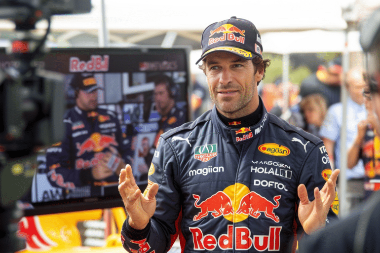 A Red Bull Formula 1 driver giving an interview in front of a camera crew. He is wearing a full Red Bull racing suit and cap, gesturing with his hands while speaking. Behind him, a monitor displays a live feed of the interview, and the scene is surrounded by media and team personnel.