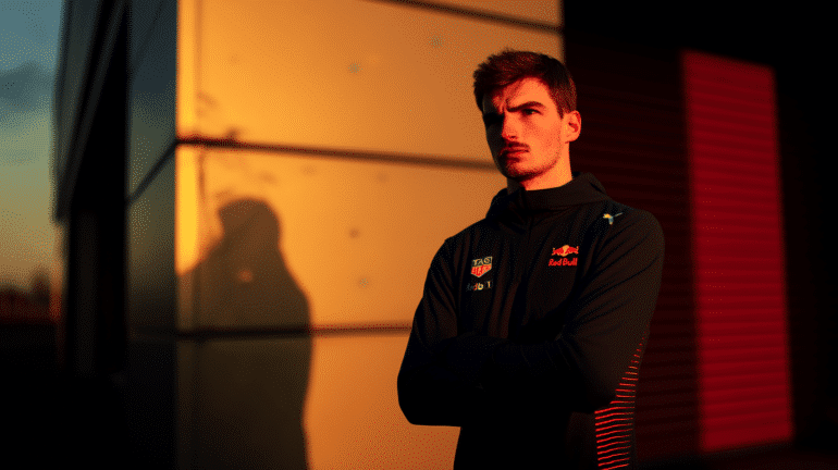 Formula 1 driver standing in front of the Red Bull Racing headquarters at sunset with arms crossed, in a serious and reflective mood.