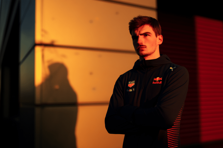 Formula 1 driver standing in front of the Red Bull Racing headquarters at sunset with arms crossed, in a serious and reflective mood.