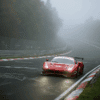 A Ferrari GT3 race car powers through a tight corner on a foggy, damp Nürburgring Nordschleife during a secret test session. The red car’s headlights pierce the mist, with no crowd present — only wet track, graffiti-covered tarmac, and quiet forest surroundings creating a focused and stealthy testing atmosphere.