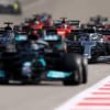 A tight group of Formula 1 cars battles down a straight section of the track, with Mercedes, Red Bull, and Ferrari cars visible. The image captures the intensity of pack racing, with heat distortion in the background and sharp tire marks along the red-and-white curbs. ​