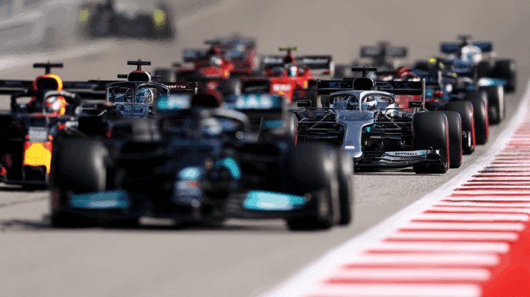 A tight group of Formula 1 cars battles down a straight section of the track, with Mercedes, Red Bull, and Ferrari cars visible. The image captures the intensity of pack racing, with heat distortion in the background and sharp tire marks along the red-and-white curbs. ​