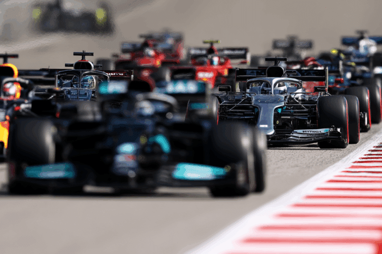 A tight group of Formula 1 cars battles down a straight section of the track, with Mercedes, Red Bull, and Ferrari cars visible. The image captures the intensity of pack racing, with heat distortion in the background and sharp tire marks along the red-and-white curbs. ​